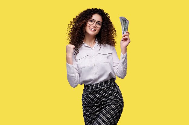 Caucasian curly hair woman holds money banknotes dollars celebrating victory and success very excited with raised arm