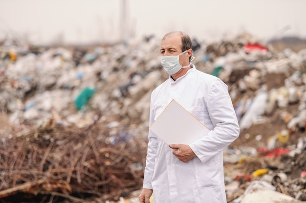 Caucasian ecologist in white uniform holding clipboard under armpit, walking on landfill and estimating pollution.