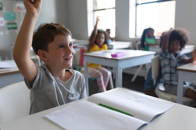 Caucasian elementary schoolboy raising hand while sitting with multiracial classmates in background