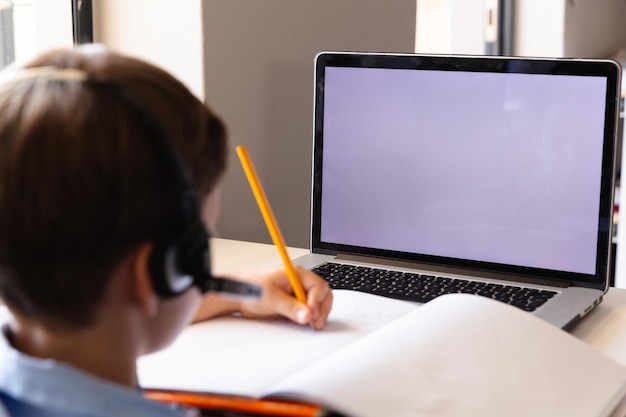 Caucasian elementary schoolboy writing on book while using laptop with copy space at desk in class