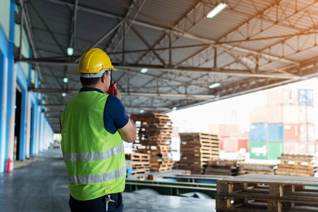 The caucasian engineer using walky talky comunicate with\
lifting container team with blue sky logistics transportation\
industry forklift handling container box loading stacker back side\
worker view