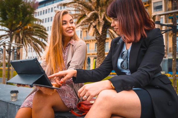 Caucasian entrepreneur session young blonde in raincoat with flower dress and brunette girl with black jacket on break from work Looking closely at the tablet
