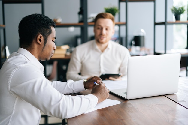 Caucasian European young businessman explaining to African coworker business plan sitting at desk