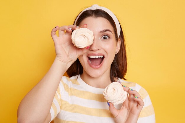 Caucasian excited woman holds marshmallows in hands wearing striped shirt and glasses covering her eye with sugary sweets looking at camera with open mouth posing isolated over yellow background