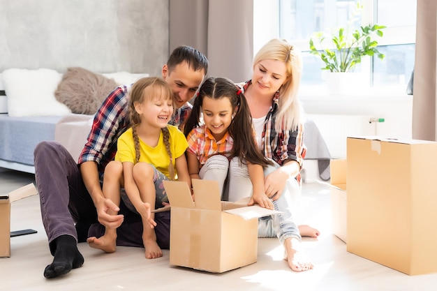 Caucasian family, man, woman and two girls sit on floor, unpack boxes and smile in new house. Behind them moving boxes.