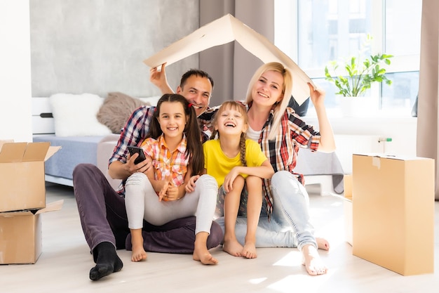 caucasian family, man, woman and two girls sit on floor, unpack boxes and smile in new house. behind them moving boxes.