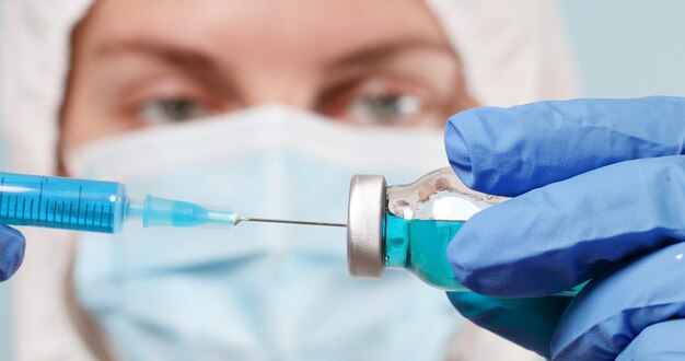 Caucasian female doctor in protective gloves and mask fill syringe with medicine for injection