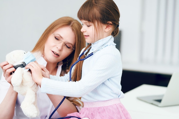 Medico femminile caucasico con bambina paziente che esamina con lo stetoscopio orso giocattolo in ufficio