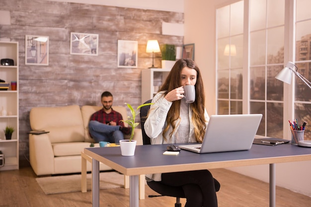 Caucasian female enjoying a cup of coffee while working on laptop from home. Boyfriend browsing on phone while relaxing on sofa.