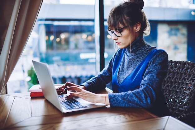 Libera professionista caucasica con occhiali ottici che lavora in remoto con un dispositivo portatile in caffetteria