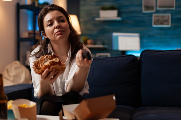 원격 시청 코미디 시리즈를 사용하여 채널을 변경하는 손에 맛있는 햄버거를 들고 백인 여성