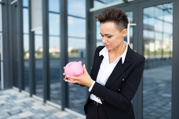 Caucasian female in office black and white clothes keeps a pink pig moneybox and waits a coleague near the office building