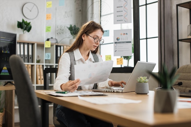 Caucasian female office worker in eyeglasses and white shirt talking and showing financial report