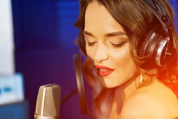 Caucasian female singer with headphones and closed eyes sings at the mic in a recording studio.