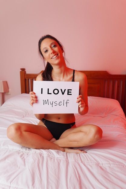 Caucasian female sitting on a bed in lingerie and holding a white paper with 'I love myself' text