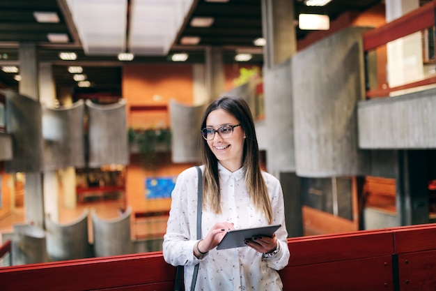 Caucasian female student standing in collage building hall and holding tablet.