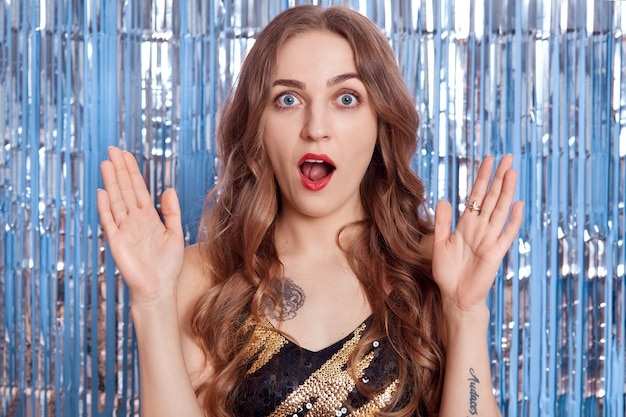 Caucasian female wearing black dress and looking at camera with widely open mouth surprised with news, posing against blue wall