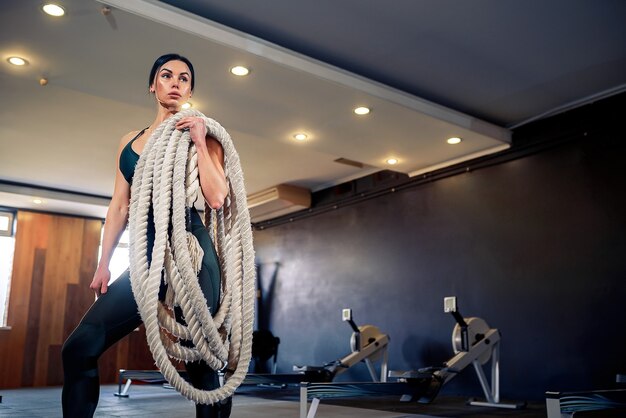 La donna in forma caucasica si è vestita in abbigliamento sportivo in posa con corde di battaglia in palestra