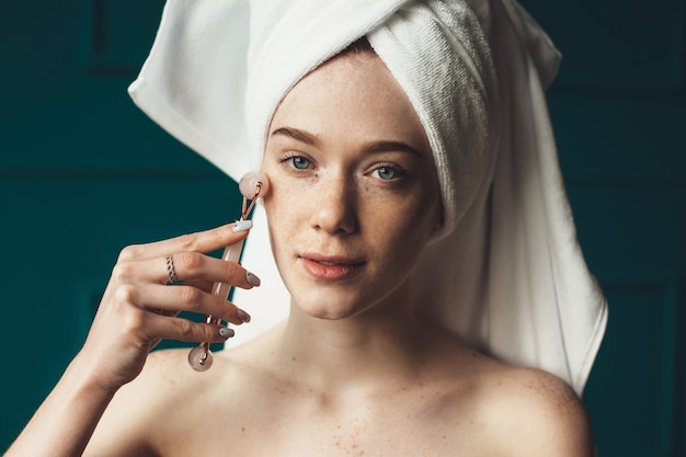 Caucasian ginger woman with freckles is massaging her face with a derma roller looking at camera with bare shoulders