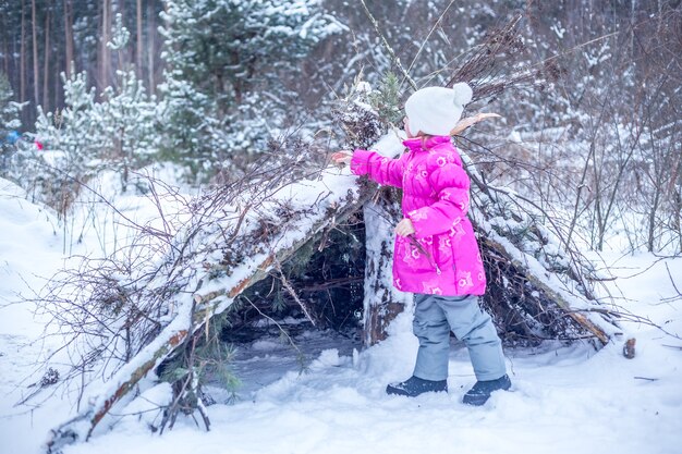 Caucasian girl of 5 years builds a hut from coniferous branches in the winter forest, playing outdoors in winter