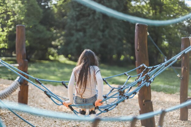 Caucasian girl climbs a rope swing in the park on the playground from the back