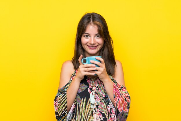 Caucasian girl in colorful dress over isolated yellow wall holding hot cup of coffee