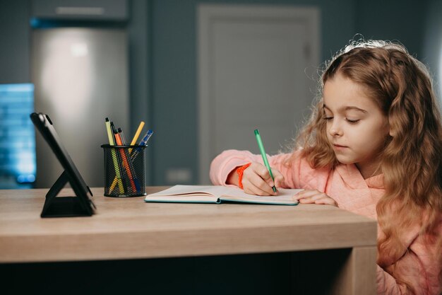 Caucasian girl doing homework seated at desk in her room listening audio lesson on tablet elearning ...