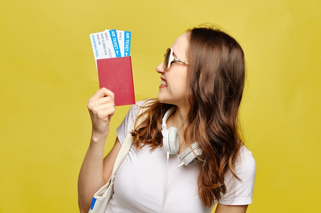 A caucasian girl in glasses looks at a passport with airline tickets on a yellow background as a vacation concept.