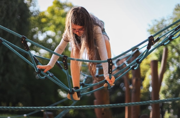 A caucasian girl halfsitting shorts climbs on a rope swing in a park on a playground