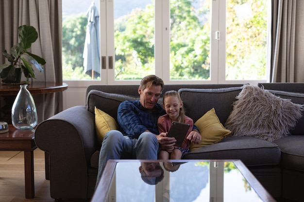 Caucasian girl and her grandfather enjoying their time in a house together, sitting on a couch, embracing, using a digital tablet