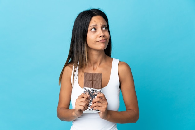 Caucasian girl isolated on blue background taking a chocolate tablet and having doubts