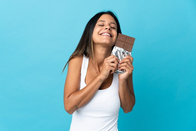 Caucasian girl isolated on blue wall taking a chocolate tablet and happy