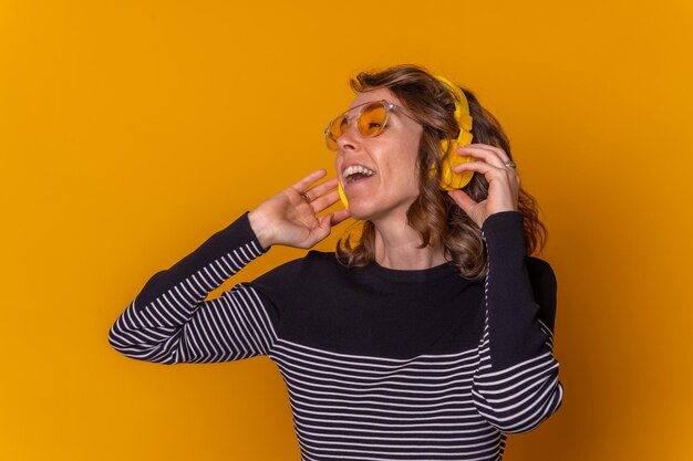 Caucasian girl listening to music smiling and dancing with her phone headphones and sunglasses yellow background