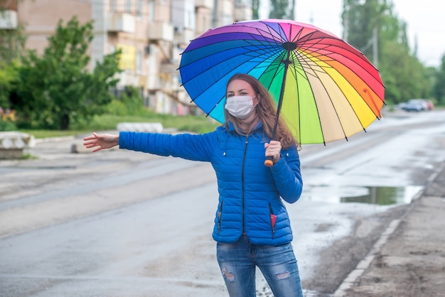 Caucasian girl in a protective mask hails taxi on an empty street, stands with an umbrella in spring rain and waits for car. Safety and social distance