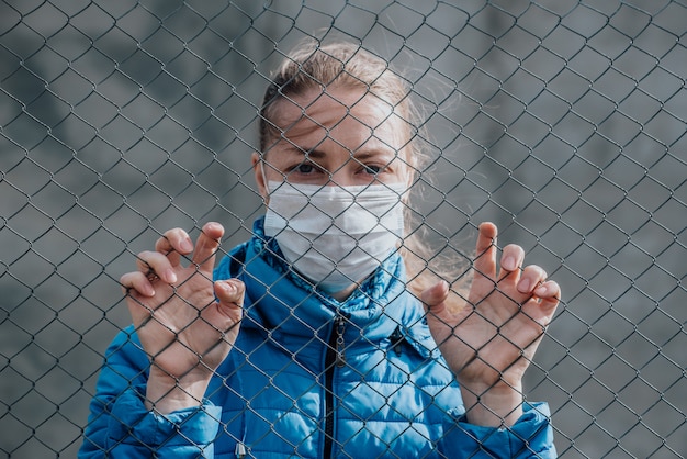 Photo caucasian girl in a protective medical mask stands behind a metal fence and looks sad. strict home isolation during quarantine.