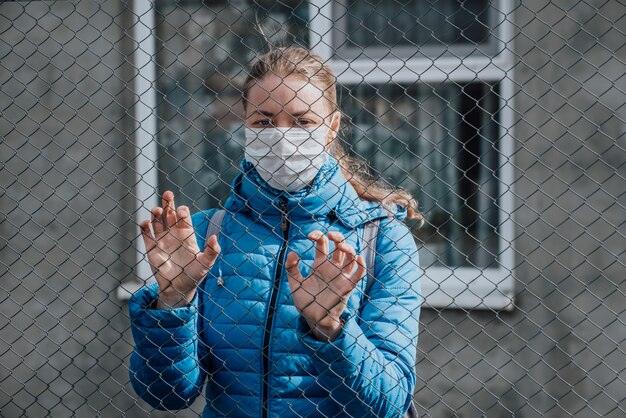 A Caucasian girl in a protective medical mask stands behind a metal fence and looks sad. Strict home isolation during quarantine.