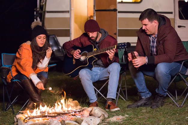 Caucasian girl roasting marshmallow over camp fire and her friend is playing on guitar with their retro camper van in the background.