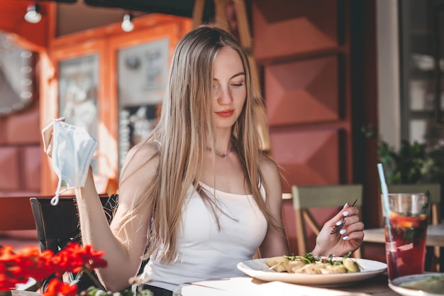 Caucasian girl sits at a table in a cafe, takes off her protective medical face mask 