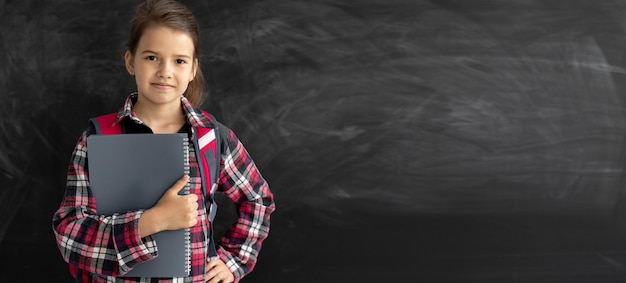 Caucasian girl student in the classroom near the chalk board he\
holds a notebook in his hands and looks at the camera online\
education and elearning concept back to school banner