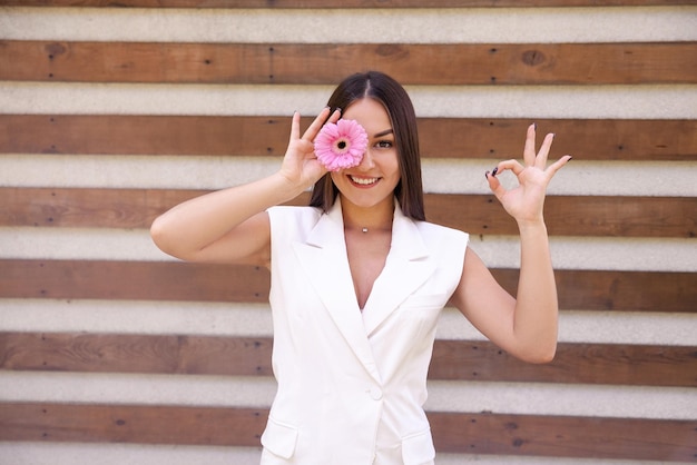 Photo caucasian girl in white clothing with a flower in front of her eye showing the okie sign