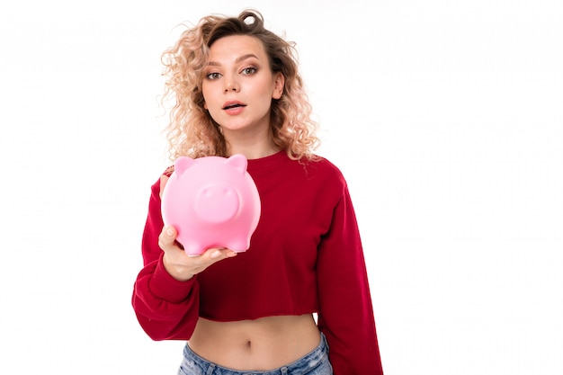 Caucasian girl with curly fair hair trying to get money from the pink pig moneybox, portrait isolated on white 