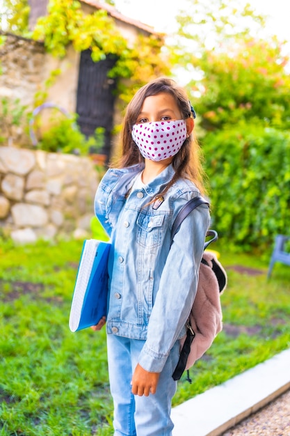 Photo caucasian girl with face mask ready to go back to school. new normality, social distance, coronavirus pandemic, covid-19. jacket, backpack, mask with pink dots and a blue block in hand