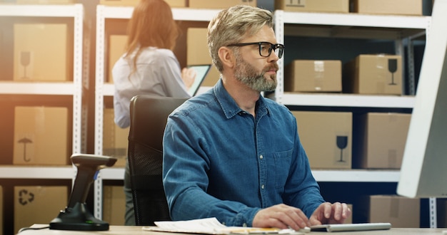 Caucasian gray-haired man in glasses working at computer screen in postal delivery store and typing on keyboard.