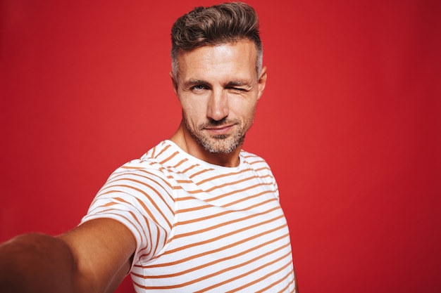 Caucasian guy 30s in striped t-shirt smiling while taking selfie photo isolated on red