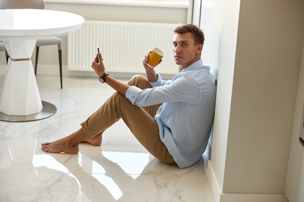 Caucasian hadsome young man in blue shirt sitting on the floor and chilling at modern kitchen