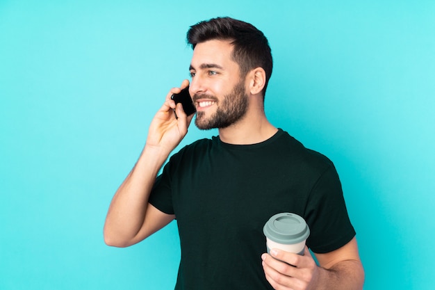 Uomo bello caucasico sulla parete blu che tiene caffè da portare via e un cellulare