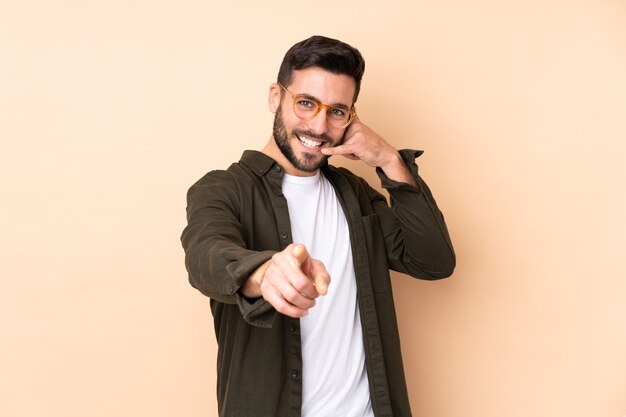 Caucasian handsome man isolated on beige wall making phone gesture and pointing front