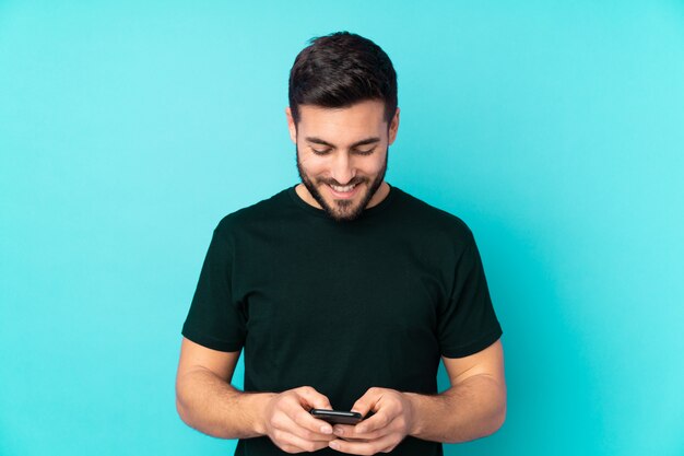 Caucasian handsome man isolated on blue wall sending a message with the mobile