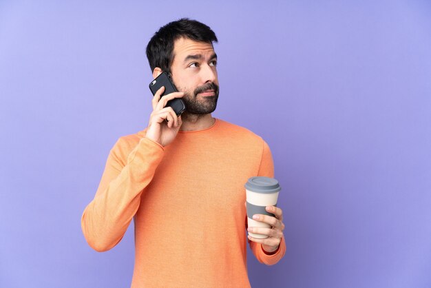 Caucasian handsome man over isolated purple wall holding coffee to take away and a mobile