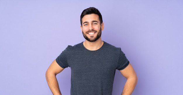 Photo caucasian handsome man posing with arms at hip and smiling over isolated purple wall